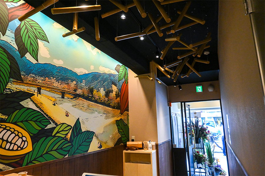 奥行きの長い京町家のような造りを生かした店内。京都の鴨川とカカオの木が壁いっぱいに描かれている。風を受けてゆらゆらと揺れる天井のウッドは、よく見ると「C」の形